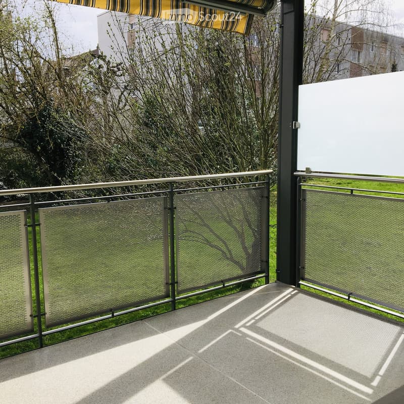 Balcony with modern metal railing, glass panels, and a concrete floor, overlooking a green garden area.