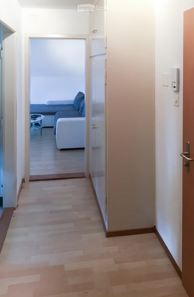 A narrow hallway with light wooden flooring and white walls, leading to a room with a sofa and coffee table visible at the end.