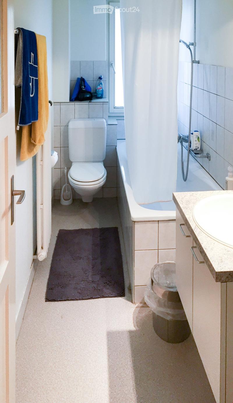 Modern bathroom with white tiles, a toilet, bathtub, sink, and a towel hanging on the door. A small rug is placed in front of the toilet.