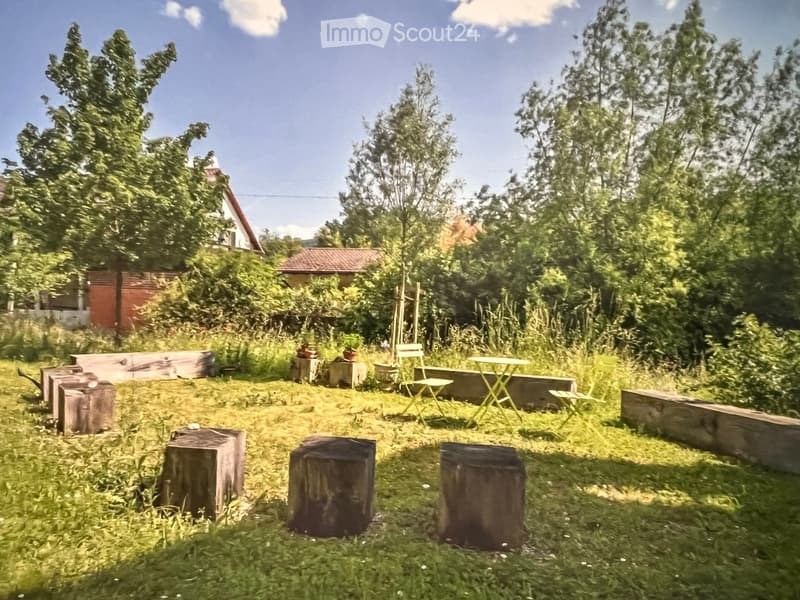 A lush garden area with wooden benches, tree stumps, and a small table and chairs, surrounded by greenery and trees.