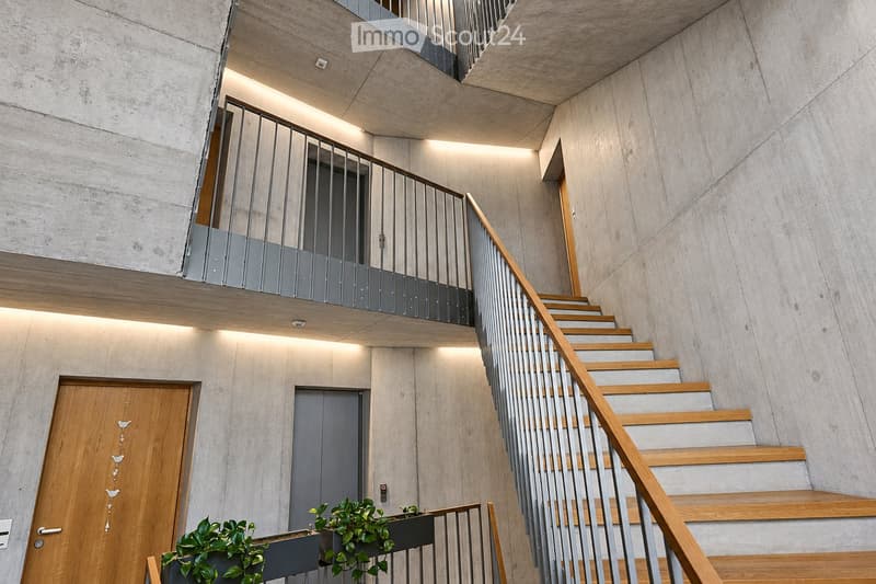 Modern interior staircase with wooden steps, metal railing, and concrete walls. Features recessed lighting and a small balcony area above.