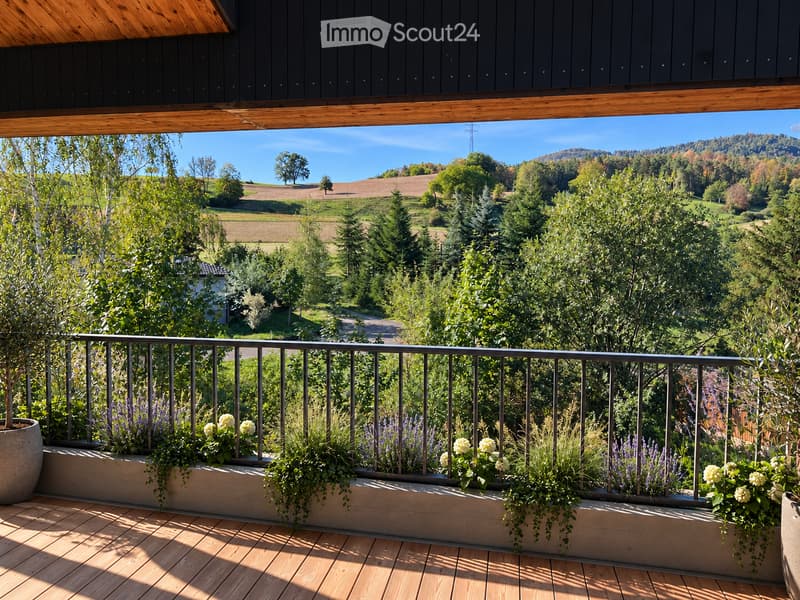 Balcony with wooden decking, metal railing, and lush greenery in planters, overlooking a scenic countryside view.