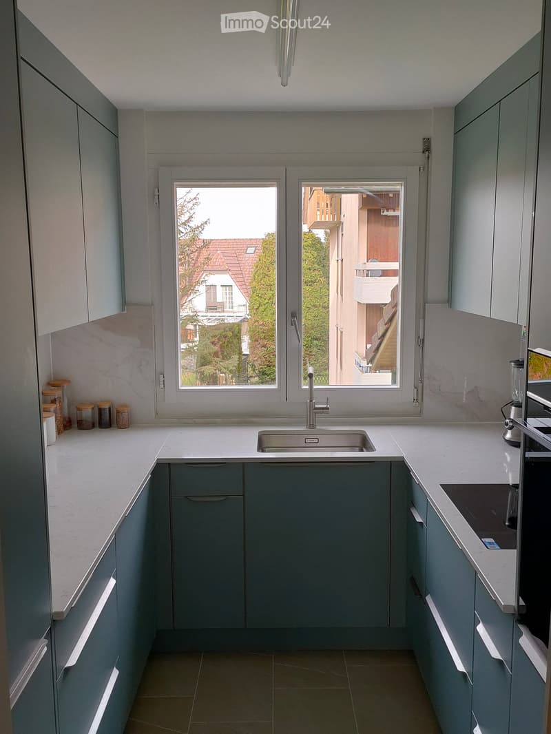 Modern kitchen with grey cabinets, white countertops, and a stainless steel sink. Features a window with a view of a balcony and outdoor area.