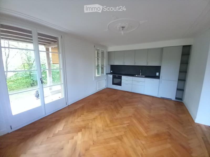 Empty room with light grey walls, wooden herringbone flooring, large sliding glass doors, and a modern kitchen area with white cabinets and stainless steel appliances.