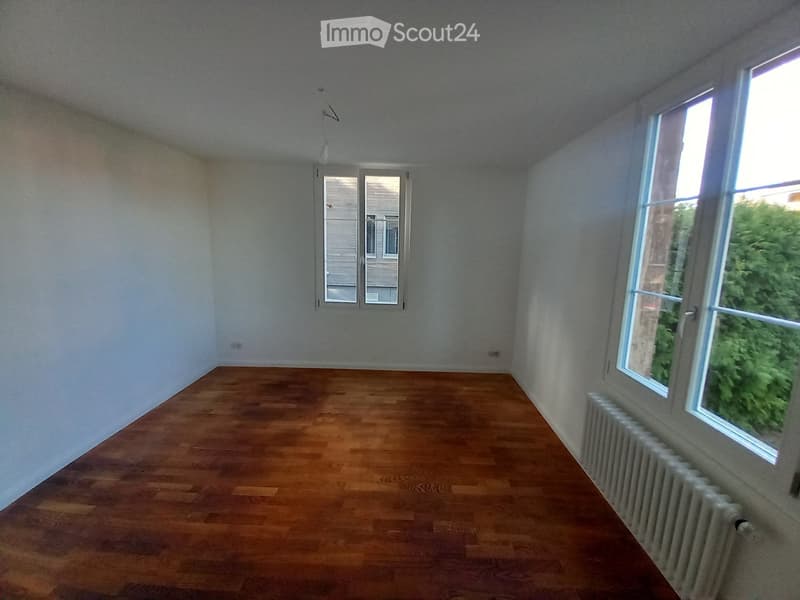 Empty room with wooden flooring, white walls, and large windows allowing natural light. The room features a radiator along one wall and a door leading to another space.