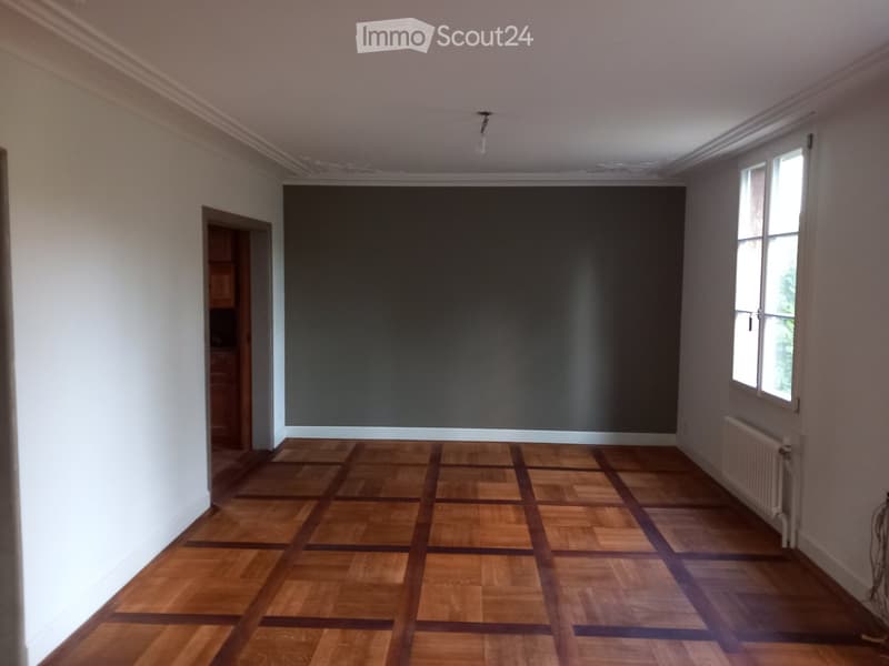 Empty room with wooden flooring, white walls, and large windows allowing natural light. There is a radiator on one wall and a doorway leading to another room.