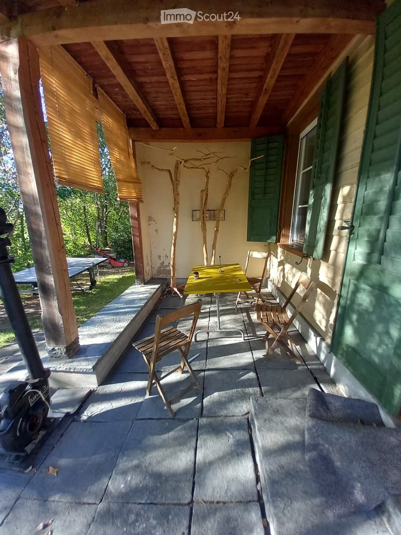 A wooden balcony with a stone floor, featuring a yellow table and several wooden chairs, with a view of a green lawn and trees.