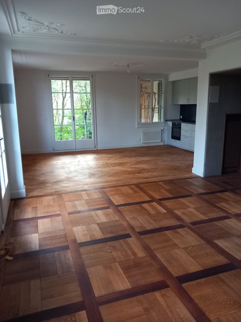 Empty room with light wooden flooring and white walls, featuring large windows and a small kitchen area with cabinets and an oven visible in the background.