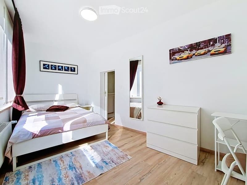 A modern, well-lit bedroom with a white bed, nightstand, dresser, mirror, and a small table with a chair. The room features wooden flooring, large windows with curtains, and artwork on the walls.