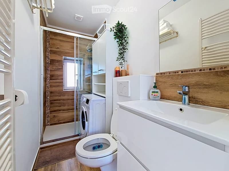 Modern bathroom with white walls, wooden accents, a toilet, sink, mirror, cabinet, washing machine, and a glass-enclosed shower with wooden paneling.