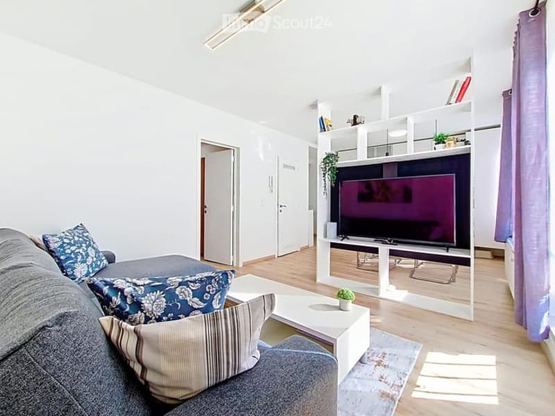 Modern living room with a grey sofa, blue and beige throw pillows, a white coffee table, a flat-screen TV on a white stand, and a small potted plant on the coffee table. The room features light wooden flooring and white walls.