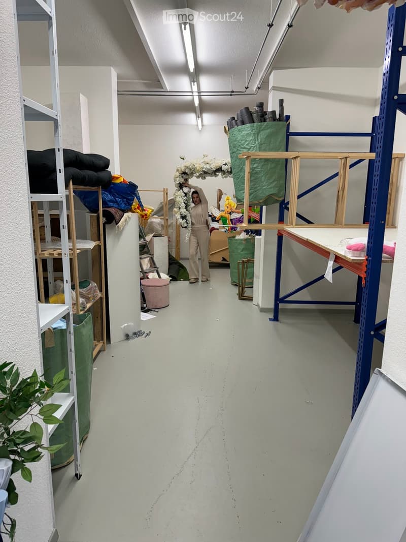 A long, empty corridor with white walls and a grey floor, featuring storage shelves and scattered items like fabric, pillows, and a floral arrangement.