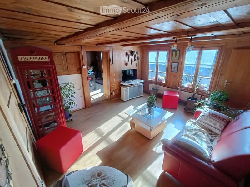 Cozy living room with wooden beams, exposed wooden ceiling, large windows, red telephone booth, sofa, coffee table, ottoman, TV, and plants.