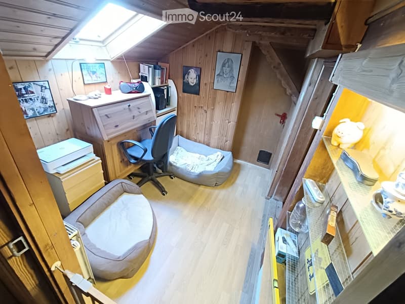 A small, cozy room with wooden walls and a skylight, featuring a desk with a laptop, a chair, a cat bed, a small stool, and framed pictures on the walls.
