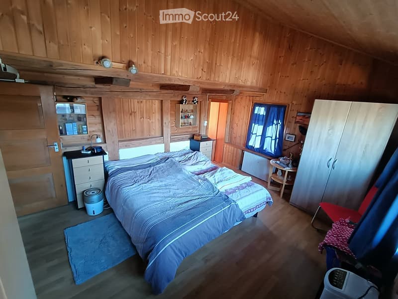 Cozy wooden bedroom with a large bed, nightstand, wardrobe, and a small table with a lamp. The room features wooden walls and a slanted ceiling, with a window covered by blue curtains.