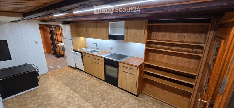 Modern kitchen with wooden cabinets, stainless steel appliances, and a central island with a sink and stove. The space features wooden shelving units and a refrigerator in the background.