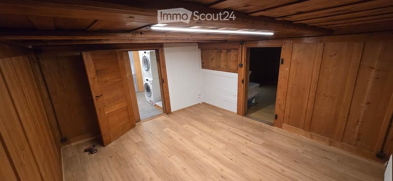 Empty room with wooden walls and floor, exposed wooden ceiling beams, and two open doors leading to adjacent spaces.