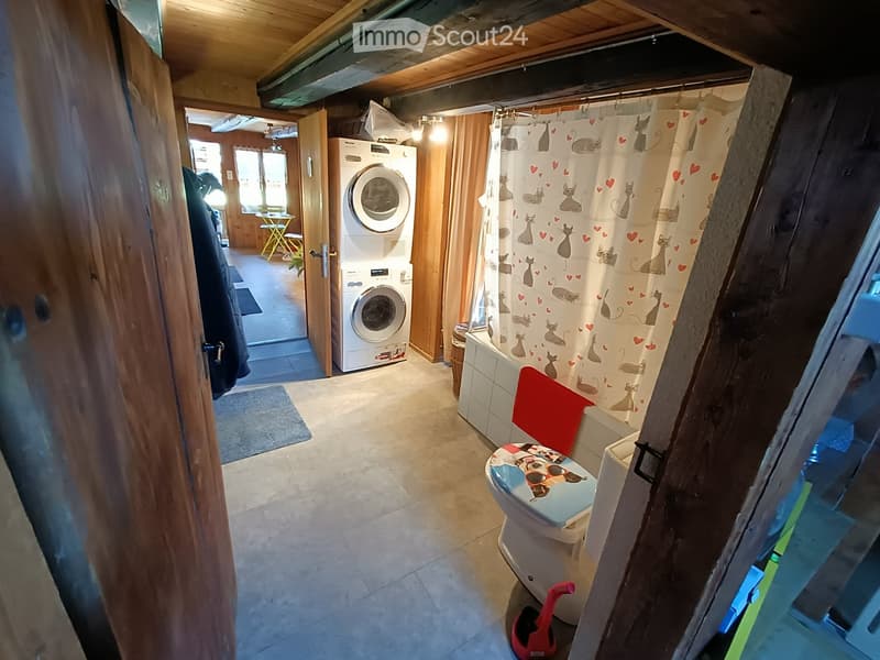 Bathroom with a toilet, bathtub, shower curtain, washing machine, and a small stool. The space has wooden walls and a tiled floor.