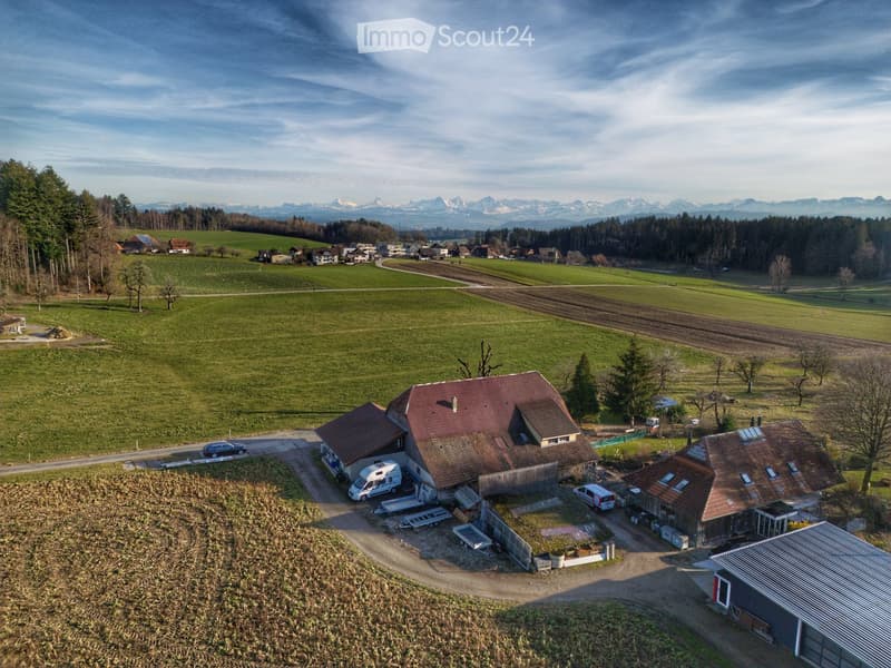 Aerial view of a rural house with a red-tiled roof, surrounded by fields, trees, and a few parked vehicles.