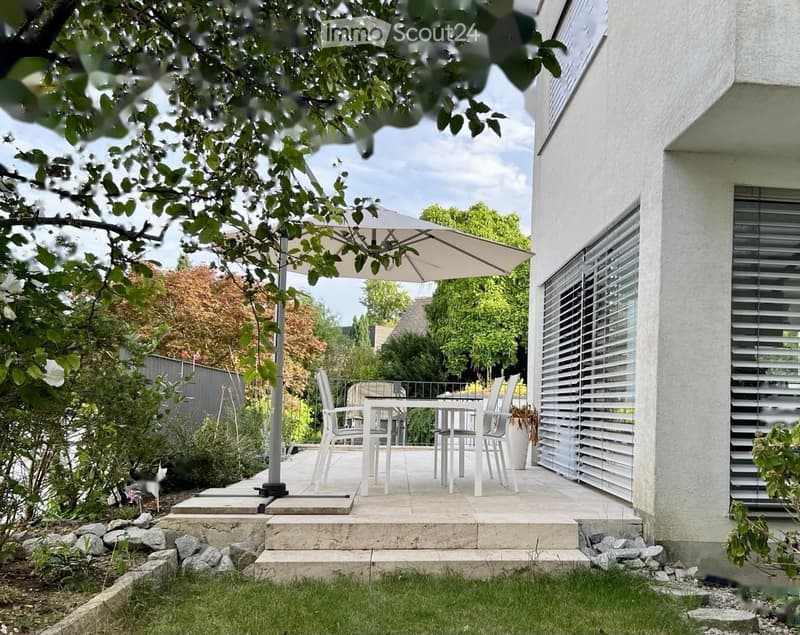 A modern outdoor patio area with a white dining set, umbrella, and surrounding greenery.