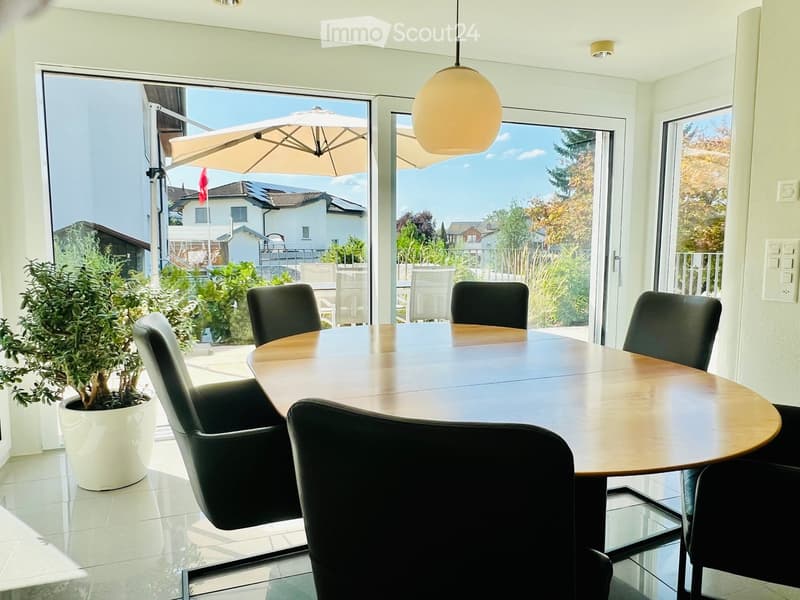 Bright dining room with a round wooden table and black chairs, large windows with a view of the garden and outdoor seating area.