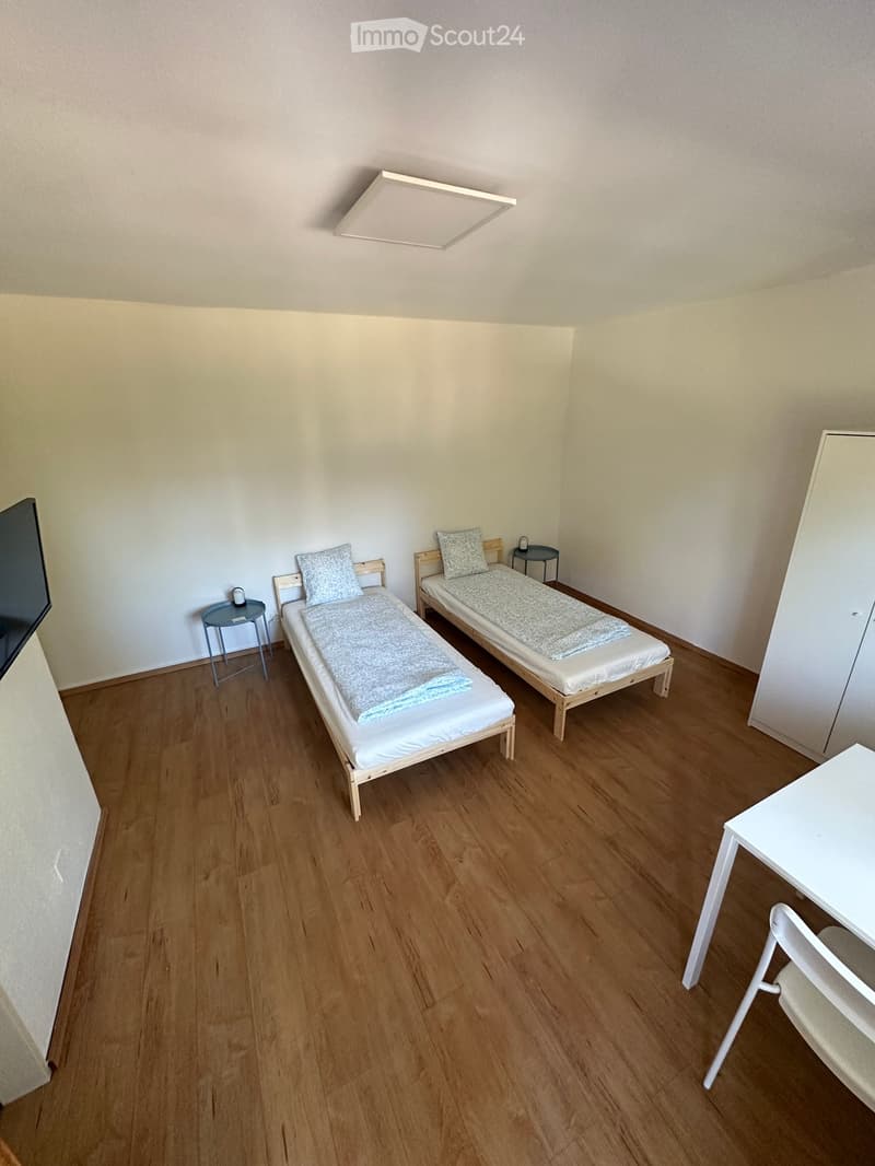 Small furnished room with two single beds, a small table, a chair, and a nightstand.