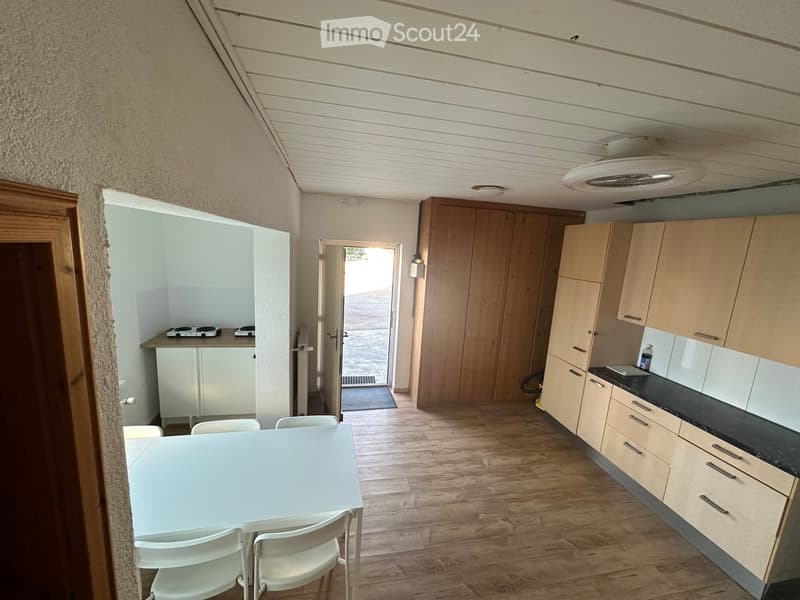 The image shows a modern kitchen and dining area with light wooden cabinets, a black countertop, a white dining table with four chairs, and a small kitchenette area with a stove and refrigerator. The floor is made of light wood, and there is a door leadin