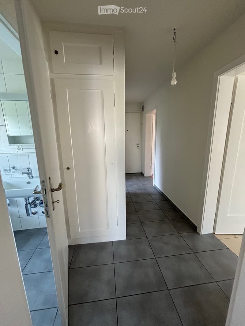 White walls, tiled floor, sink, mirror, dark grey tiles, and an open door leading to another room.