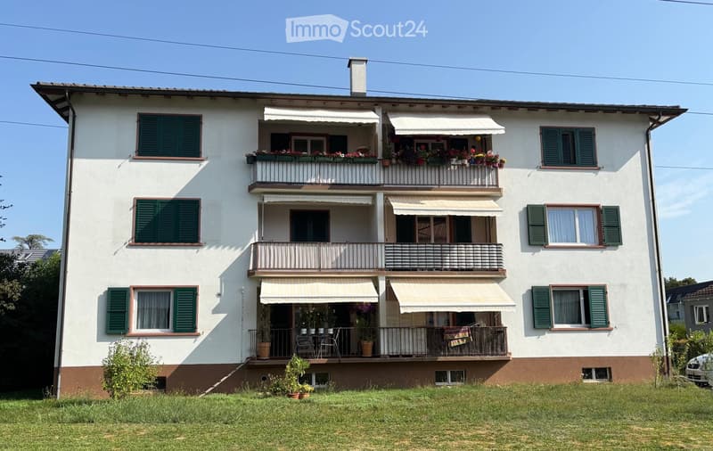 4 story building, white and brown exterior, green shutters, covered balconies, flowering plants on balconies, small garden in front