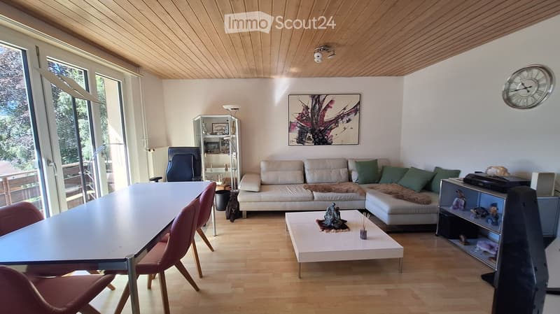 Bright living room with wooden floors, white walls, and large windows. Features a cream-colored sofa with green cushions, a coffee table with a decorative item, a dining table with chairs, a floor lamp, and a picture frame on the wall.