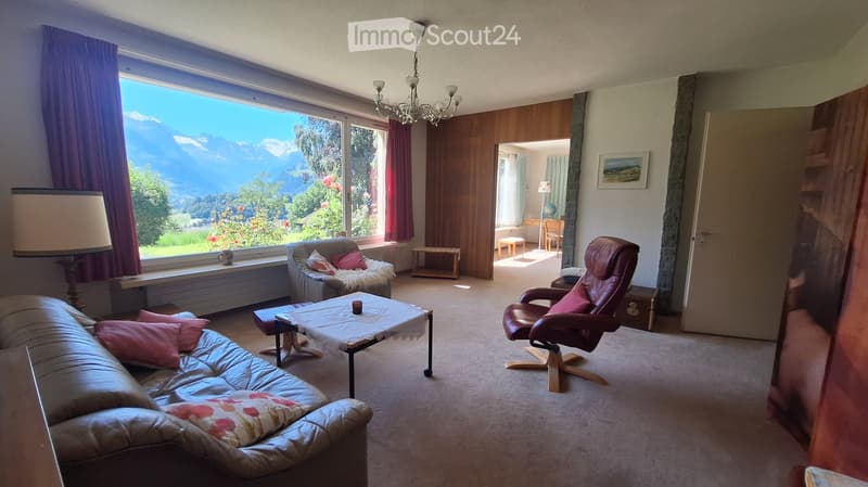 Bright living room with large windows offering a scenic mountain view, furnished with a sofa, coffee table, armchair, and a small table with a lamp.