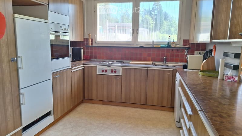 A modern kitchen with wooden cabinets, a stainless steel refrigerator, oven, and sink. The countertop is made of wood, and there are various kitchen appliances and items visible, including a toaster, cutting board, and dish soap.
