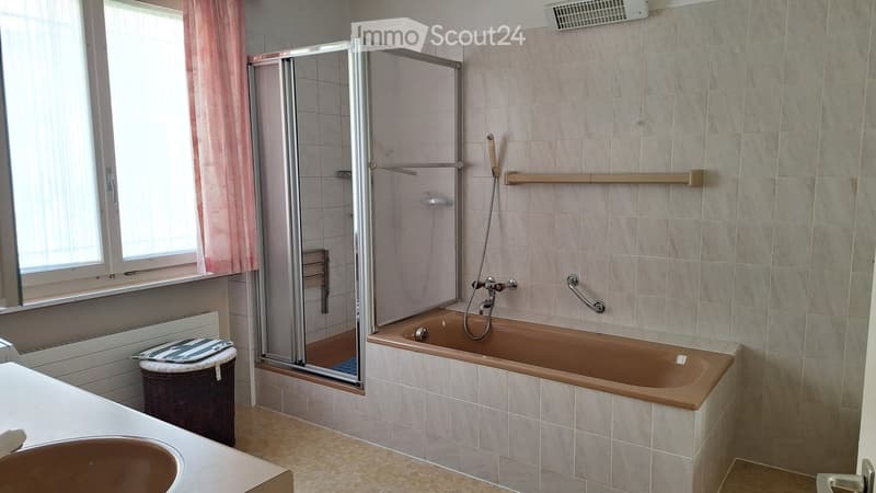 Bathroom with a bathtub, shower, sink, radiator, and a window with a curtain. Includes a handrail and a shelf in the shower area.