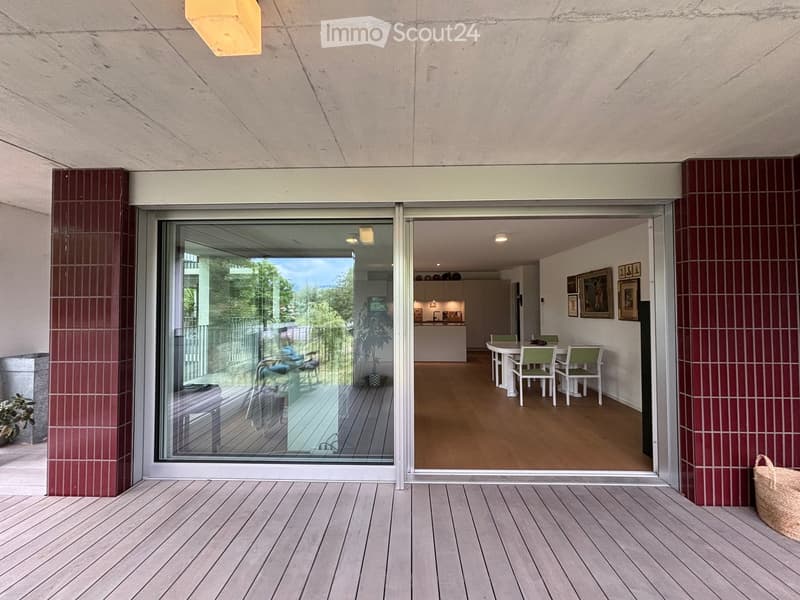 Modern balcony with wooden decking, large glass sliding doors leading to a dining area inside, featuring a round table and chairs, with a view of greenery outside.