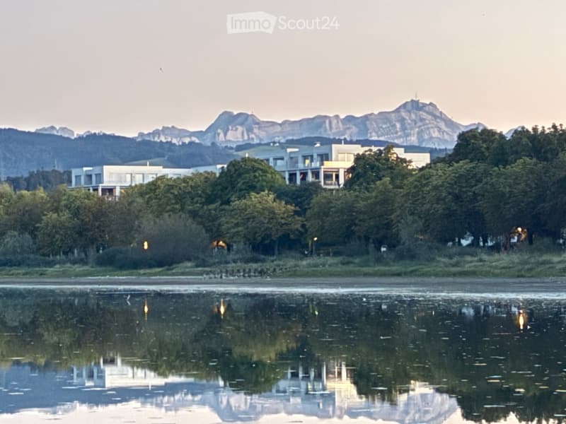 A serene outdoor scene with a calm body of water reflecting the surrounding landscape, including modern buildings and distant mountains.