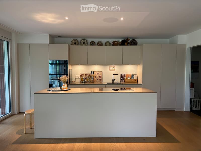 Modern kitchen with a white island, light wood flooring, and grey cabinets. Features a sink, decorative plates, and a vase with dried flowers on the island.