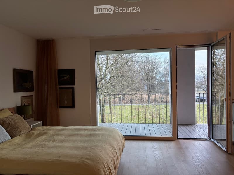 Bright bedroom with wooden flooring, large bed, nightstand with lamp, framed pictures on wall, and sliding glass doors leading to a balcony with metal railing and outdoor view.