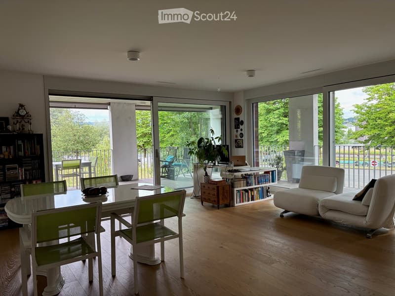 Modern living and dining area with large glass doors leading to a balcony, featuring a white sofa, dining table with chairs, bookshelf, and laptop on a small table.