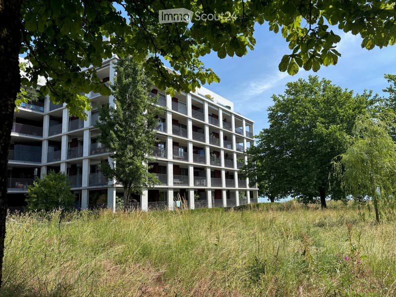 Modern multi-story residential building with balconies, surrounded by trees and grassy area.