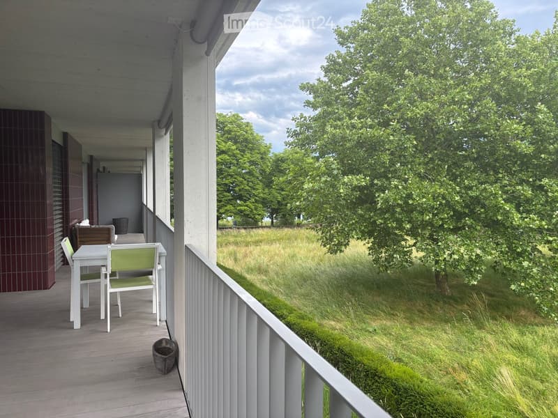 Balcony with a white table and green chairs, overlooking a grassy area with trees.