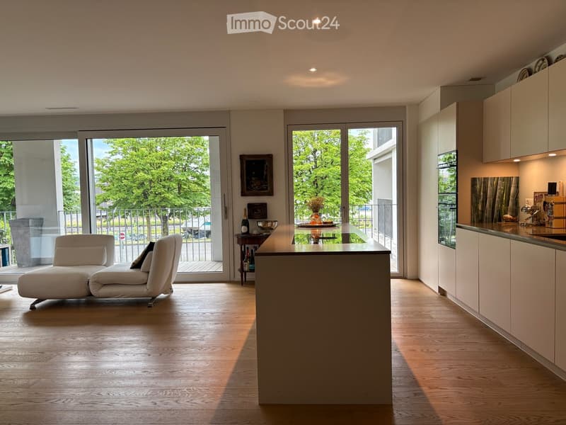 Modern open-plan living and kitchen area with light wood flooring, white cabinets, a central island, a sofa, and a small table with decorative items.