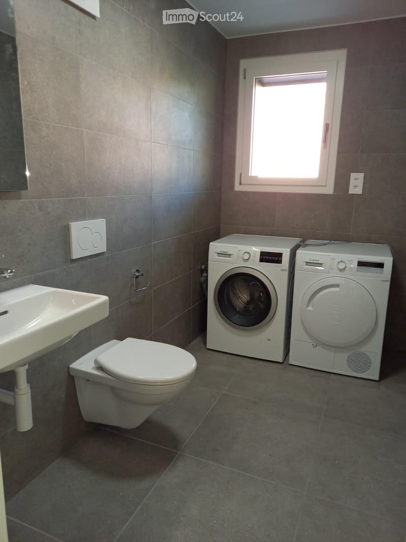 Bathroom with a toilet, sink, washing machine, and dryer, featuring tiled walls and a window.