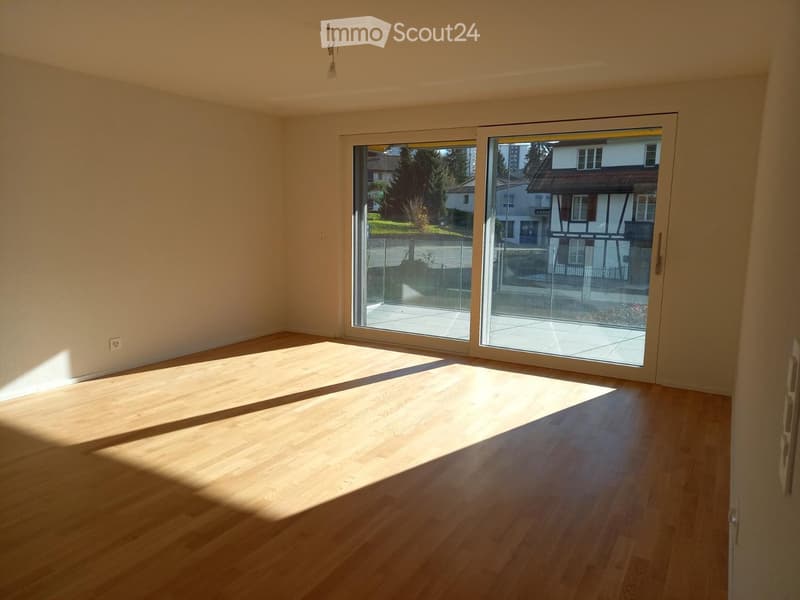 Empty room with wooden flooring and large sliding glass doors leading to a balcony or terrace. Natural light streams in, casting shadows on the floor.