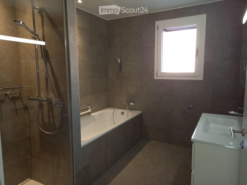 Modern bathroom with a bathtub, shower, sink, and towel rack. Features dark tiled walls and a window for natural light.