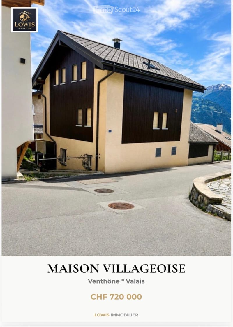 Two-story house with a combination of beige and dark wood exterior, multiple windows, and a garage entrance. The house is situated in a scenic mountainous area with a paved driveway leading to the entrance.