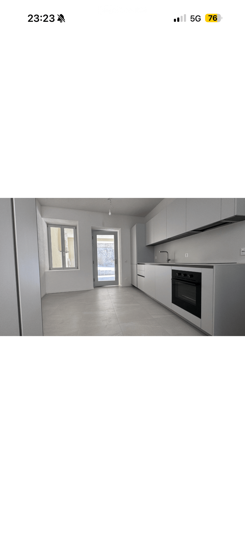 Empty kitchen with white cabinets, a sink, and an oven. The space features a tiled floor, a window, and a doorway leading to another room.