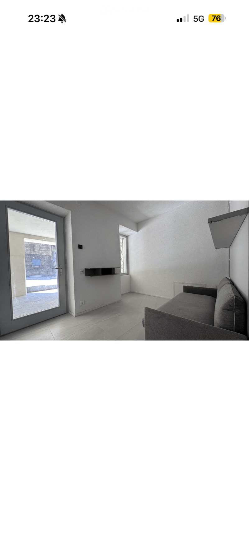 A modern, minimalist living room with a grey sofa, a sleek black shelf, and large windows offering a view of an outdoor area.