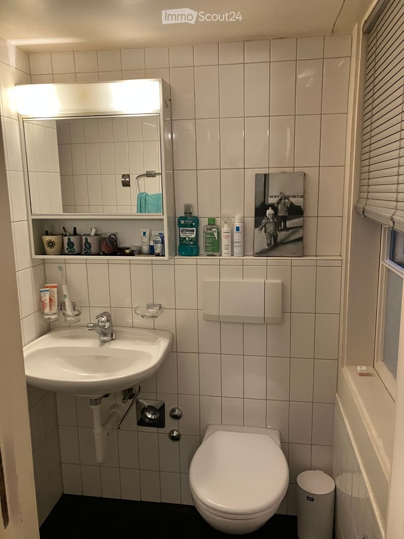Tiled walls, sink, toilet, window, shelf with toiletries, mirror, trash can