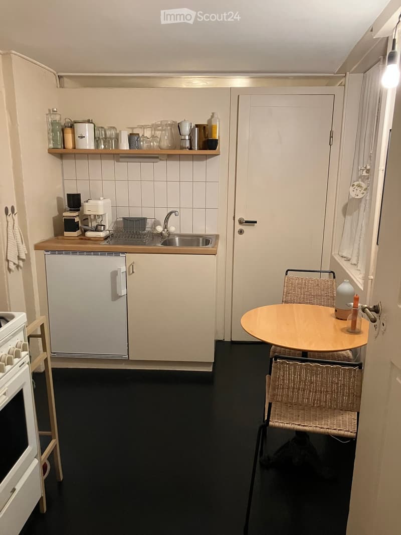 small kitchen with sink, small refrigerator, and a coffee machine. There is also a small table with 2 chairs and a stool.