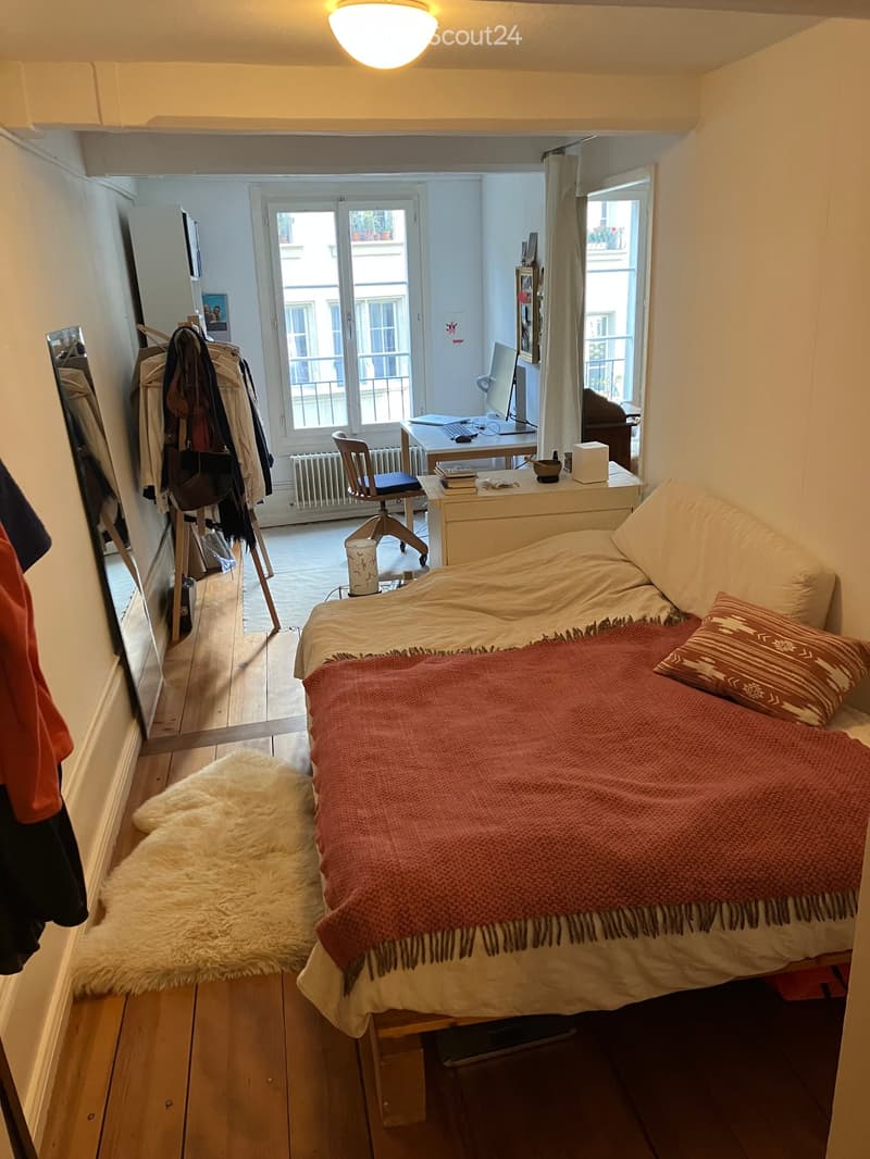 A small bedroom with wooden floor, two beds, a desk, and a radiator. Bed sheets and blankets are unmade. A mirror, closet, and clothes rack are also visible.